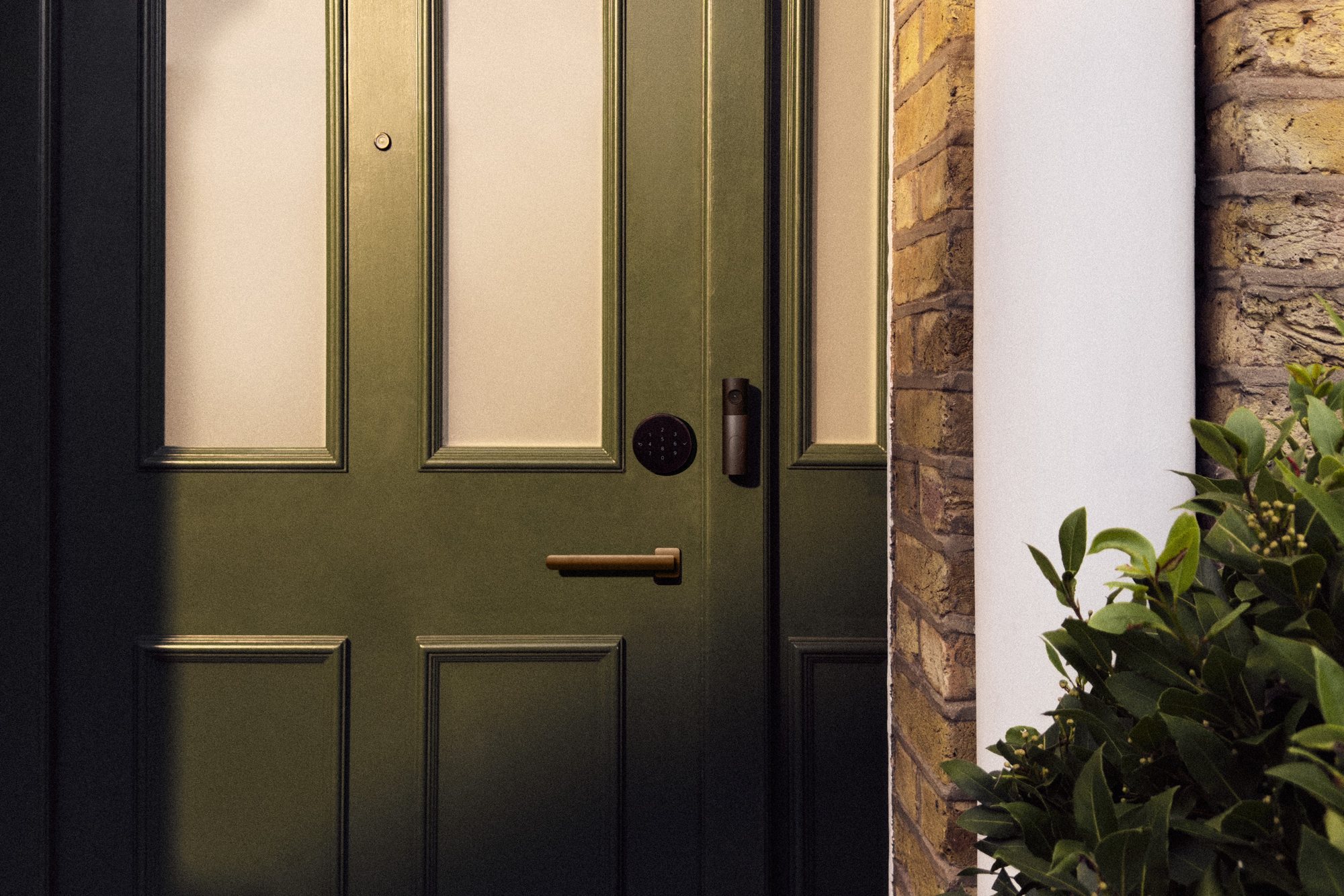 A dark green front door with a smart deadbolt, set into a brick entryway beside a leafy plant