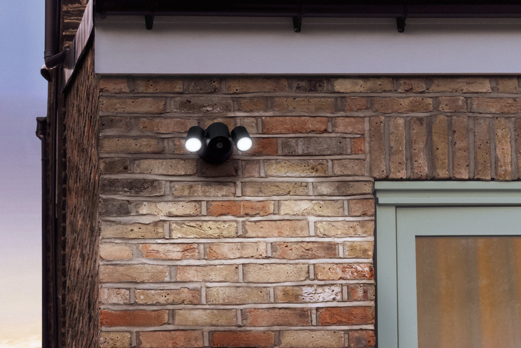 LG Floodlight Cam with two lit floodlights mounted on a brick wall above a doorway at dusk